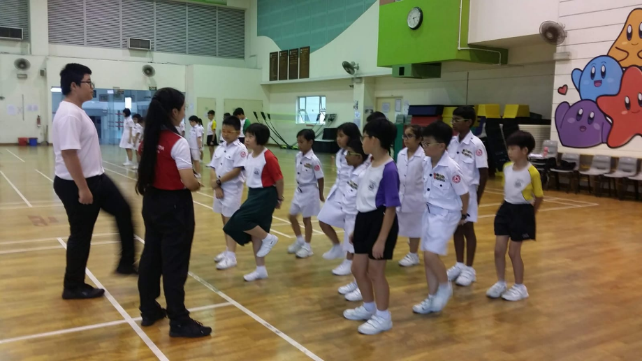 Children in white uniforms march in a line inside a school gymnasium while two adults supervise.